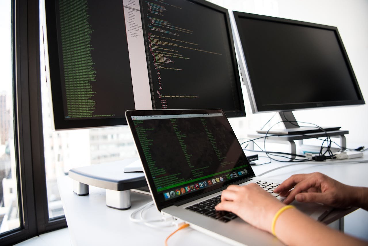 Close-up of a developer typing on a laptop with code visible on multiple monitors in the background, illustrating the technical and content work behind AI search optimization for a small business.