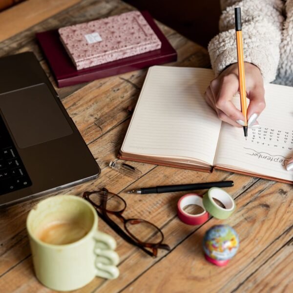 A woman planning her schedule in a notepad beside a laptop and a cup of coffee on a wooden desk