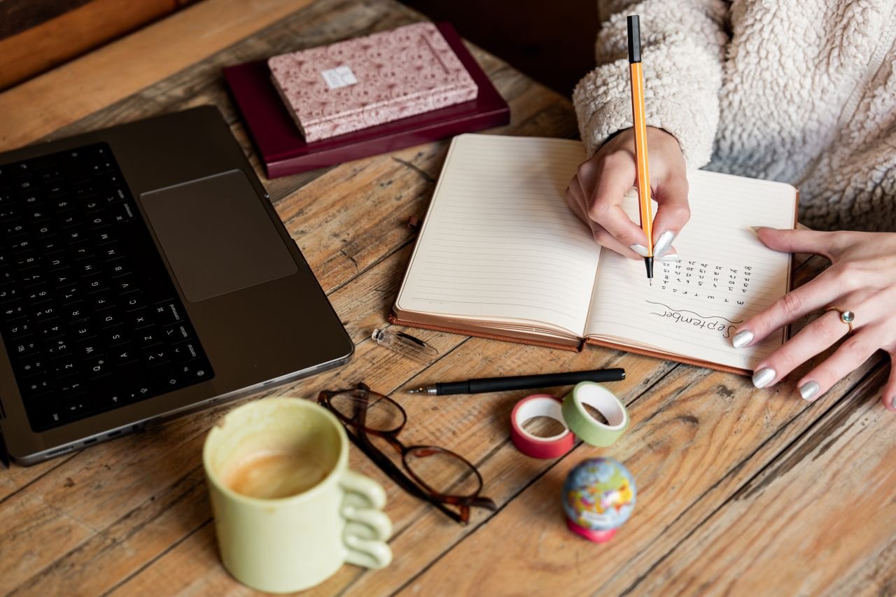 A woman planning her schedule in a notepad beside a laptop and a cup of coffee on a wooden desk