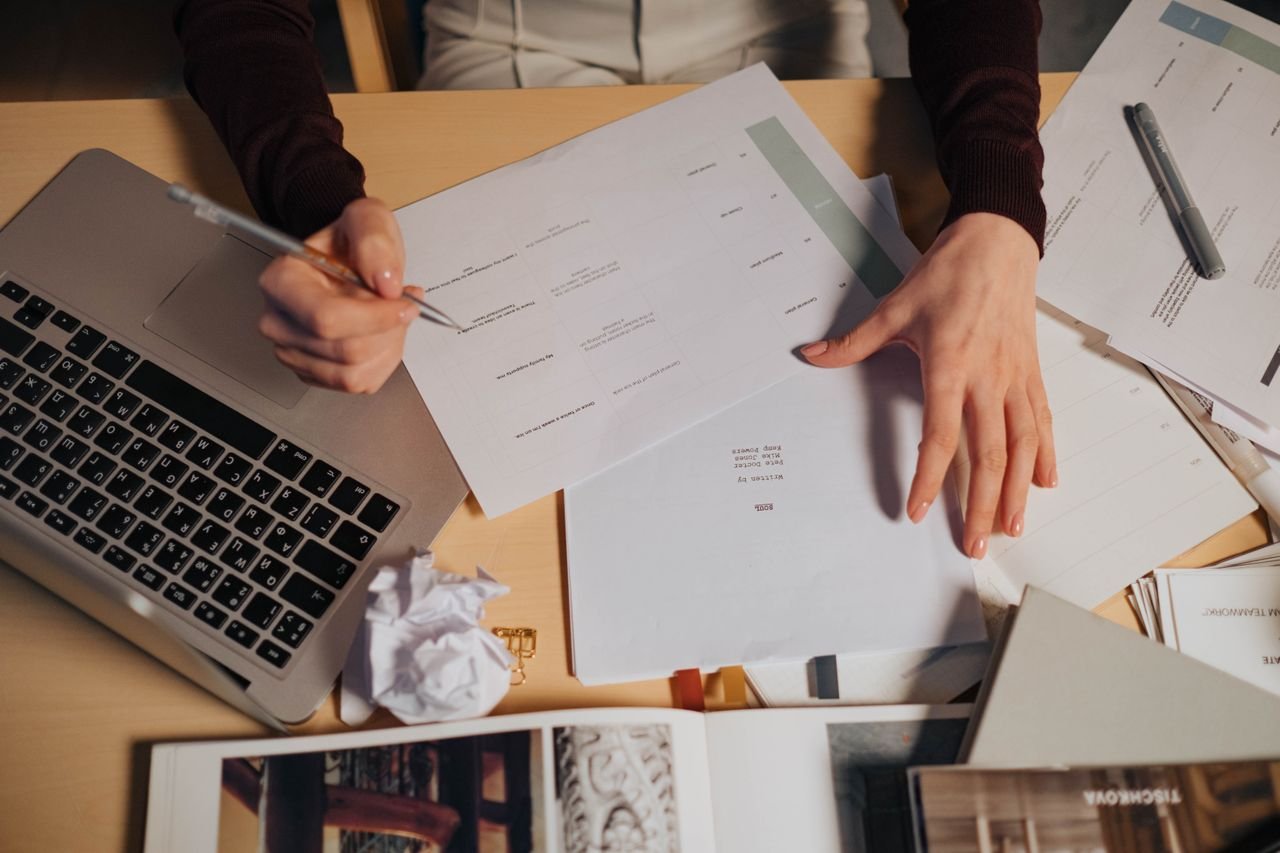 A person takes notes at a desk with a laptop and printed documents spread out in front of them...Looking at AI policies.